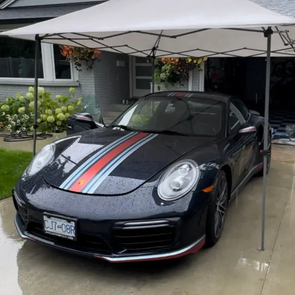 Dark blue Porsche 911 Turbo with Martini stripes under a pop-up canopy in a driveway on a rainy day